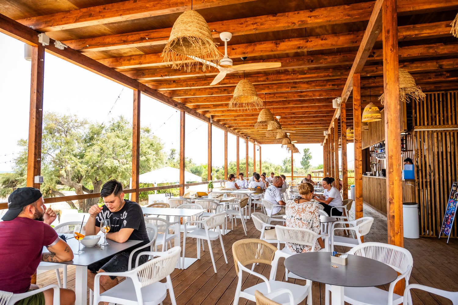 Terrazza coperta del Maka Beach piena di clienti ai tavoli, con le lampade di paglia e il bancone sul fondo